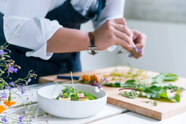 Chef en veste blanche et tablier noir prépare un plat gastronomique dans une cuisine professionnelle, saupoudrant des herbes sur une salade dans un bol blanc