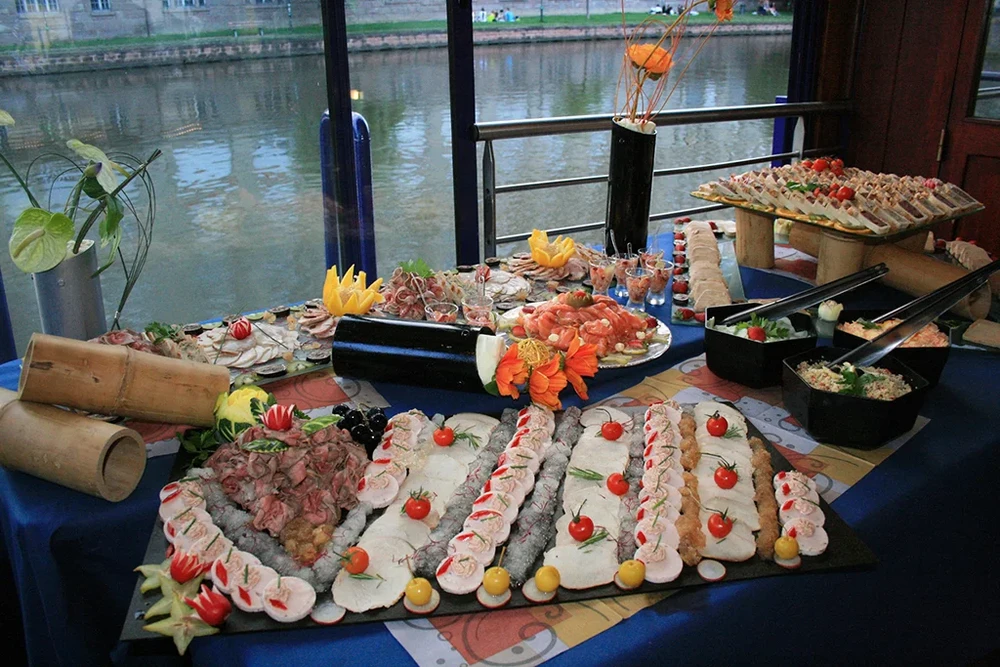 Buffet traiteur gastronomique disposé sur table bleue près d'une baie vitrée donnant sur canal urbain